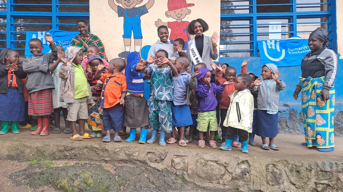 A group of children smile and wave at the camera. Behind them, Miss Jeannette also waves and smiles. They all stand in front of a school wall decorated with drawings.