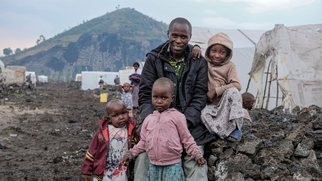 A smiling man is surrounded by three children. They are sitting on a low black stone wall. Behind them, rows of tents can be seen, and in the distance, a green hill.