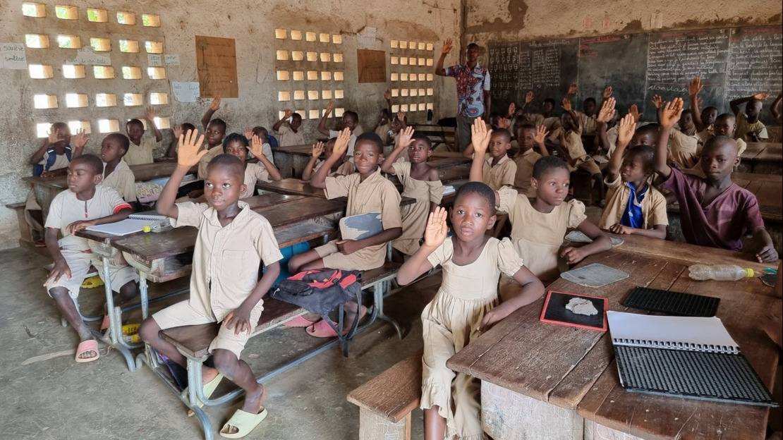 Inside a classroom. Children aged 8–10 in beige uniforms are seated around wooden desks and raising their hands.
