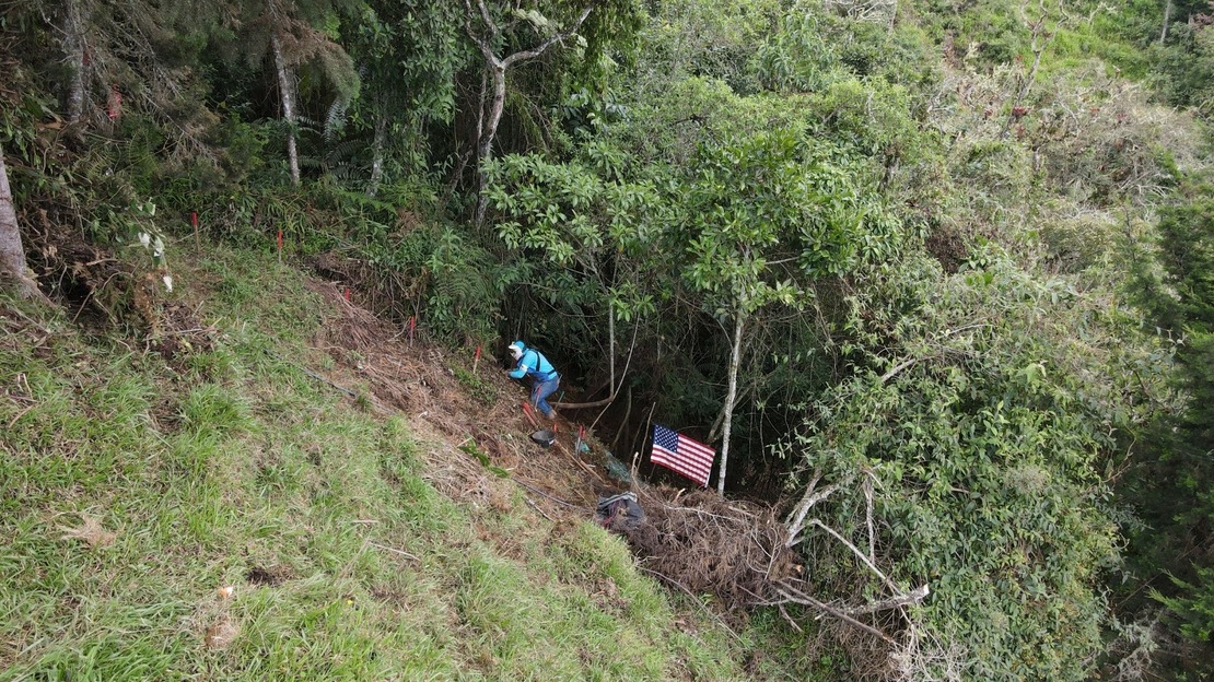 A deminer crouches on a steep slope, clearing the ground ahead of him. Around him, a lush forest. A U.S. flag stands beside him.