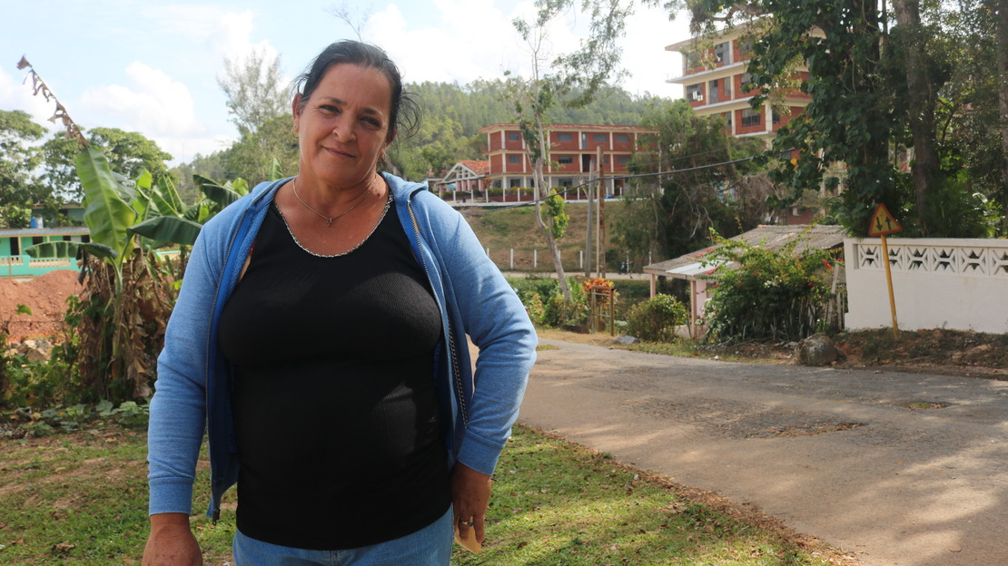 Portrait of Dania at mid-thigh, smiling. In the background we see a road, red brick houses and vegetation on the side of the road.