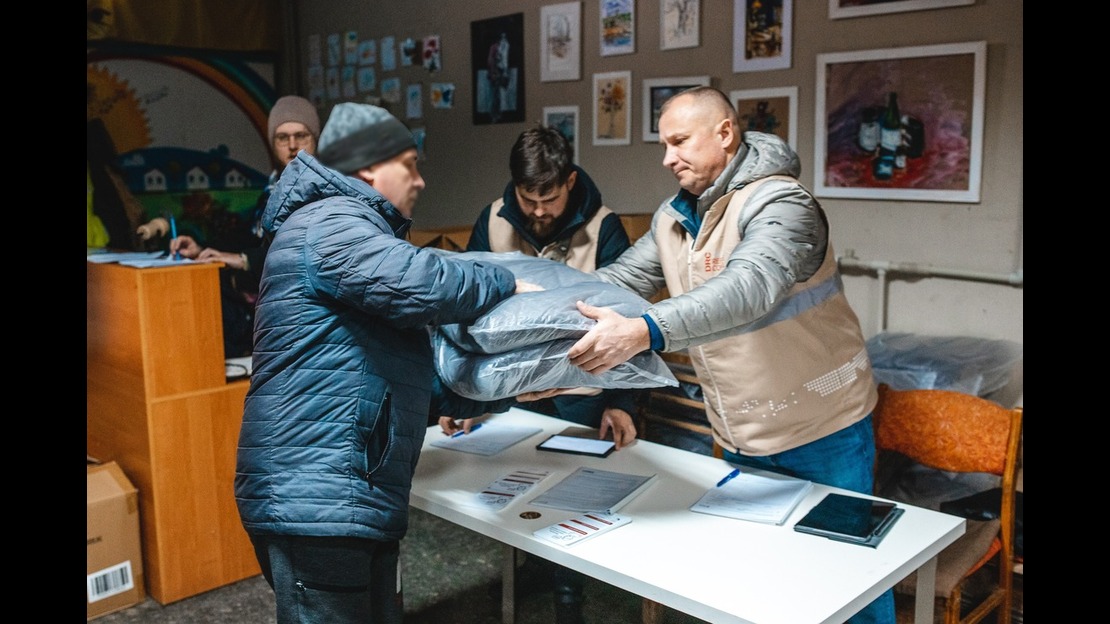 Man wearing a tan vest distributes thermal covers to another man wearing a blue coat above a white table and two men in background