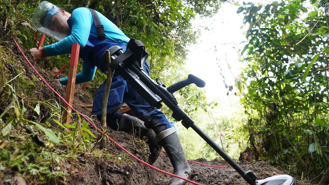 A deminer wearing protective gear, working on a muddy slope with equipment 