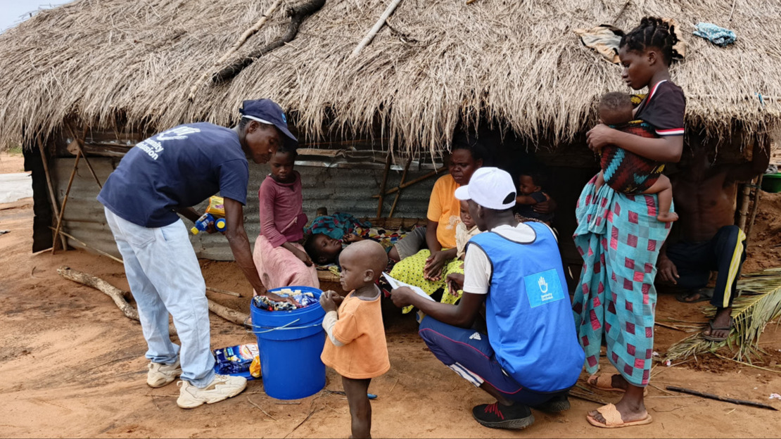 In front of a tin and straw hut, two people wearing HI t-shirts unpack the contents of a large blue bucket, showing it to a family consisting of two women and several small children.
