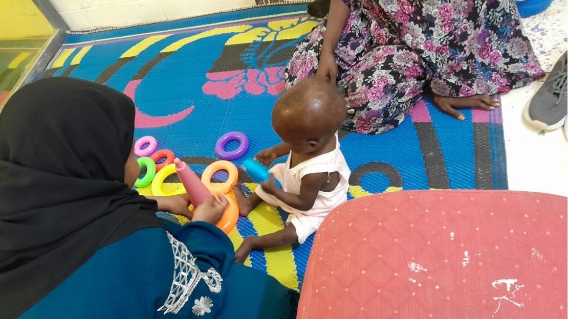 A child is sitting on a mat, holding a toy handed to him by a woman