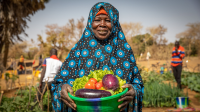 Fadima poses with a bowl of tomatoes, eggplants and lettuce in her hand. The shared garden can be seen in the background.