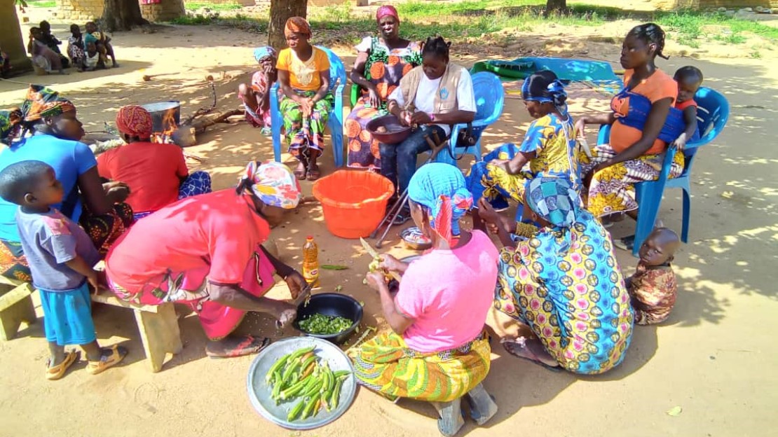 A group of women and children sit in a circle under the trees as part of a cooking activity.