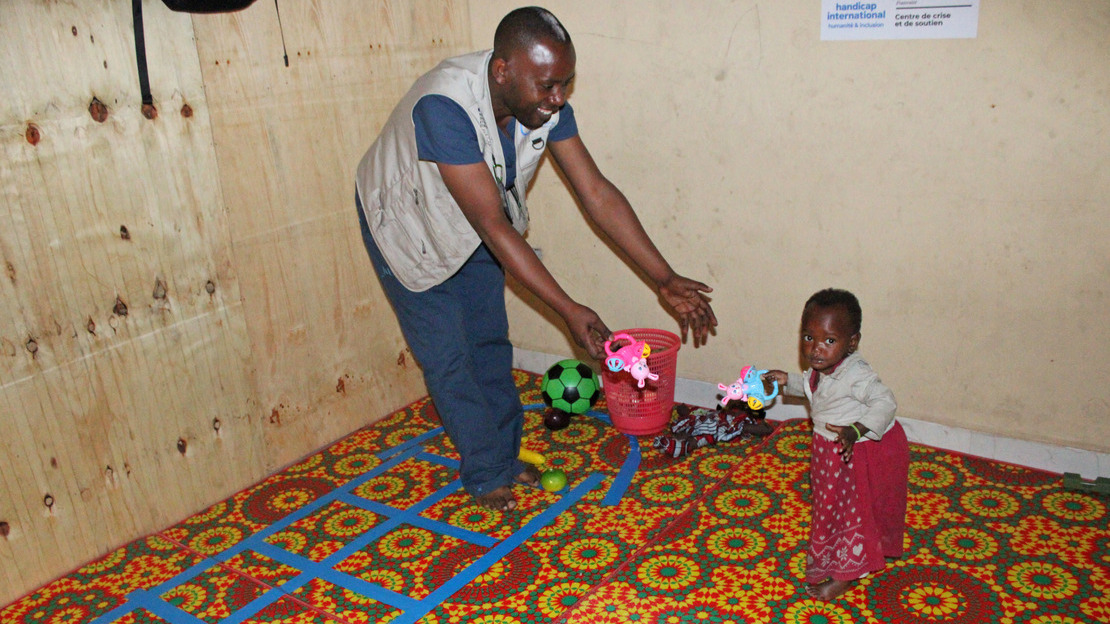A man smiles and reaches out his arms to a little girl standing in front of him, holding a plastic toy in her hand.