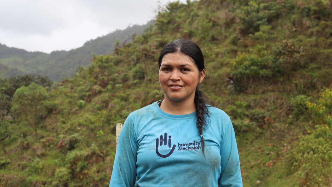 A close-up portrait of a smiling woman wearing a polo shirt featuring the HI logo and the words ‘Humanity & Inclusion’. Behind her are slopes covered in lush vegetation.