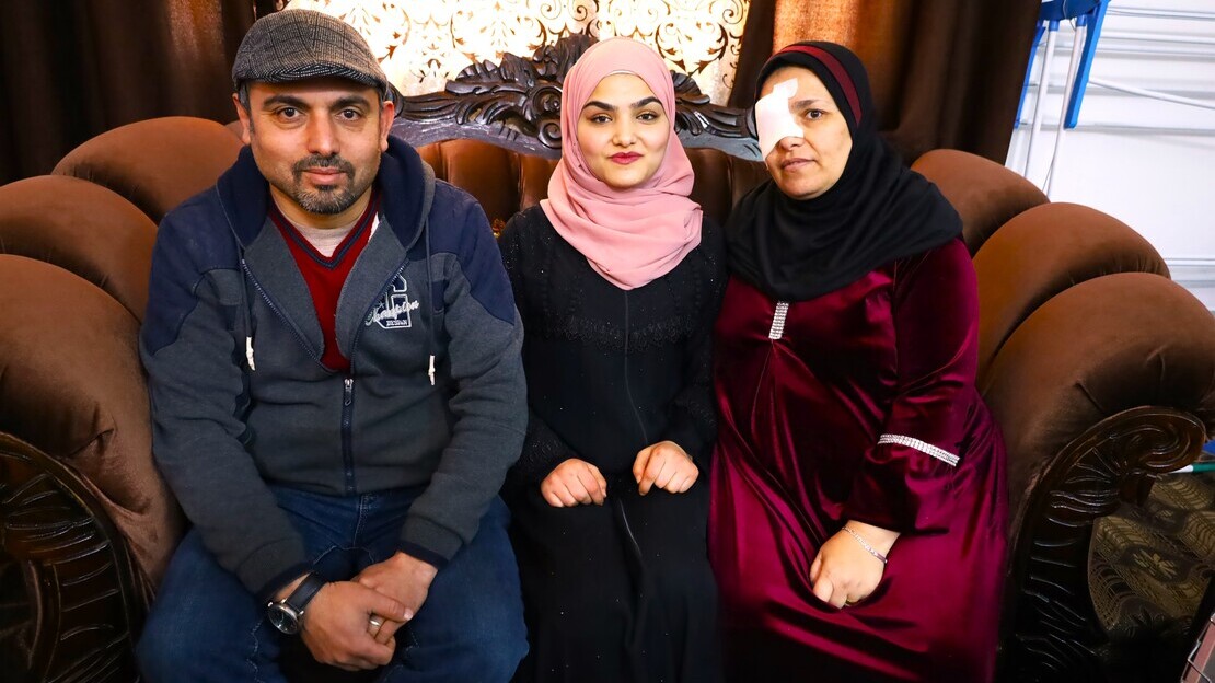 A family of three sitting on a couch. To the left, a father wearing a hat. In the middle, a young woman wearing a headcovering. To the right, a woman wearing a headcovering and eyepatch on her right eye.