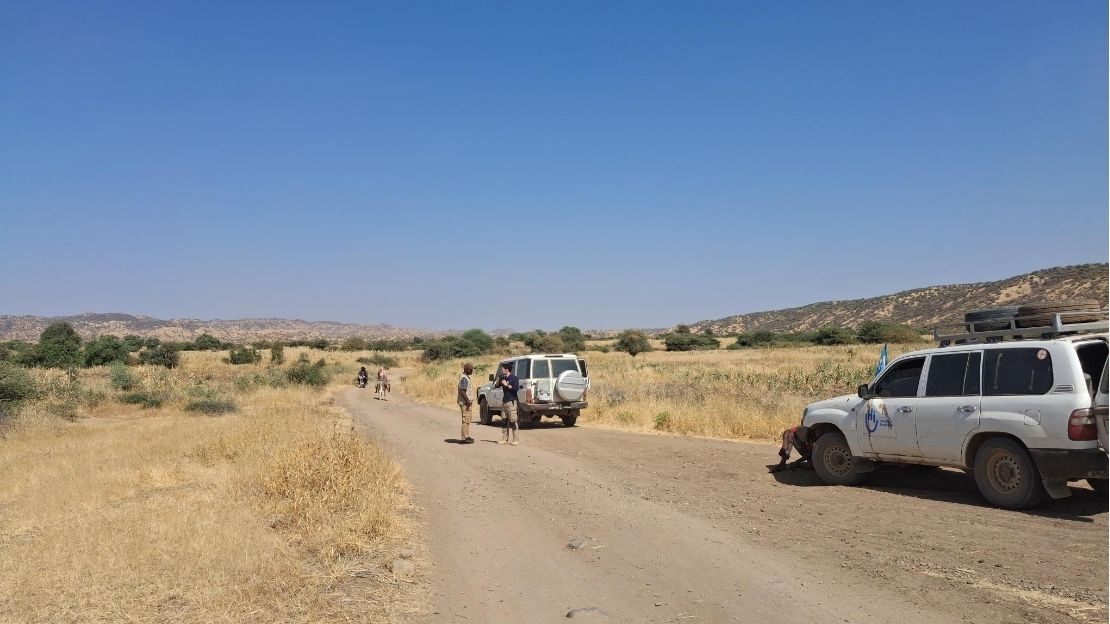 A team of four aid workers are standing and chatting on a path, near two HI vehicles.