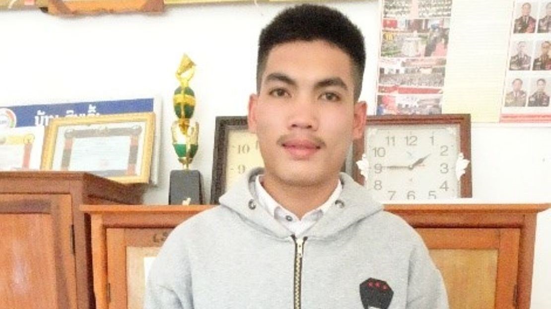 Portrait of Joc, a teenage boy, smiling looking into camera. Wearing a grey hoodie, in front of framed certificates and a clock.