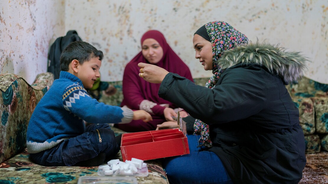 Young boy smiling, sitting on a couch and interacting with a red segmented box. He sits across a specialist, a woman wearing a floral head covering and coat with fur hood, who smiles as she interacts with the boy and activity. The boys mother, wearing a red head covering, looks on in the background from another couch, 