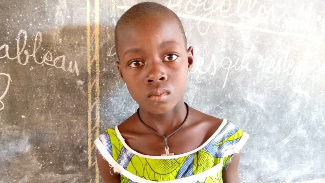 Young girl with shaved head wearing green and blue patchwork top and a necklace, standing in front of a chalkboard with faded writing 