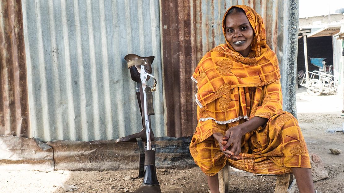 Woman wearing yellow head covering and outfit, smiling, sitting on a chair in front of a metal wall next to a prosthesis 