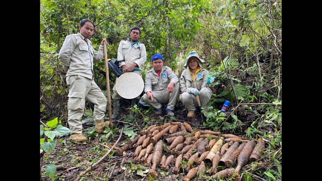 Four people are standing in an outdoor setting with dense vegetation, surrounded by trees, bushes, and branches. They are wearing light-colored work clothes and boots. Some are holding tools, including a shovel and a circular metal object. On the ground in front of the four people, a large number of long, rusty metal objects are arranged in a group. The objects vary in size but are similar in shape. A spray can is visible on the ground to the right.