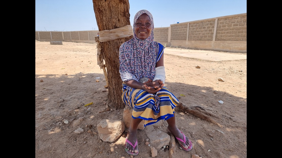 A woman is sitting on a rock at the foot of a tree in dry, sandy terrain. She is wearing a patterned top, colorful striped fabric, and sandals. A white bandage covers her right arm. In the background, a long brick wall surrounds the space under a clear sky.