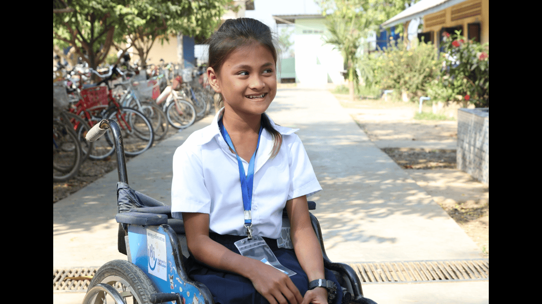 A young girl is sitting in a wheelchair with a visible logo on an outdoor path. She is wearing a white short-sleeved shirt, dark pants, and a badge hanging around her neck on a blue lanyard.  In the background, several bicycles are parked in a row along the path, under trees and near buildings. The path is lined with vegetation and a building with light-colored walls and a roof. The ground is concrete with a drainage grate visible in the foreground. 