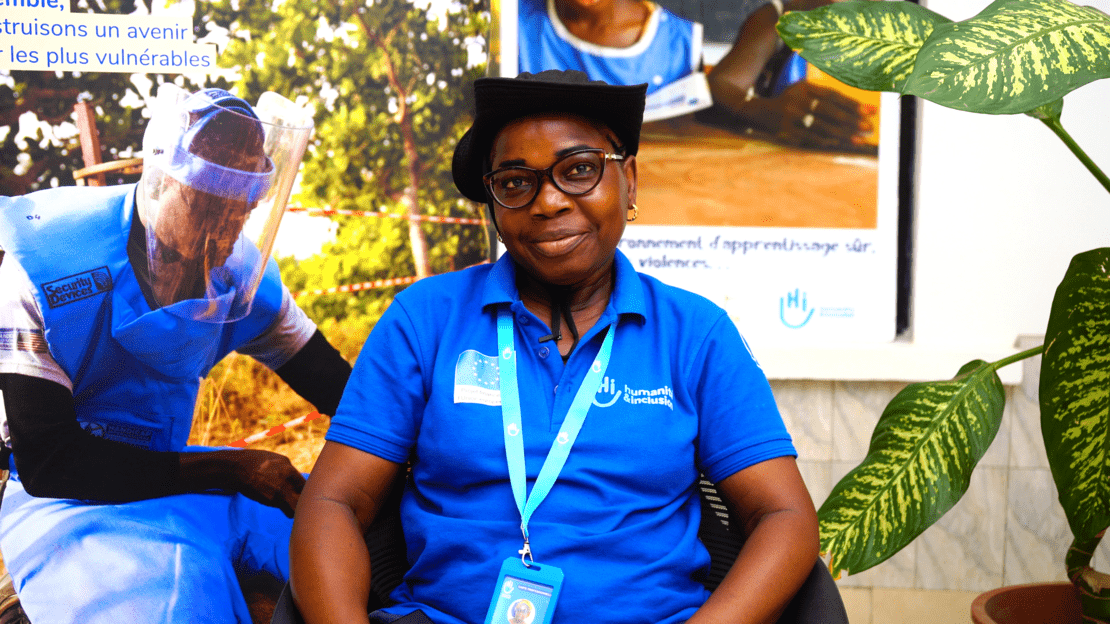 Portrait of a young woman wearing a blue HI polo and blue lanyard, sitting in front of demining posters and smiling at the camera.