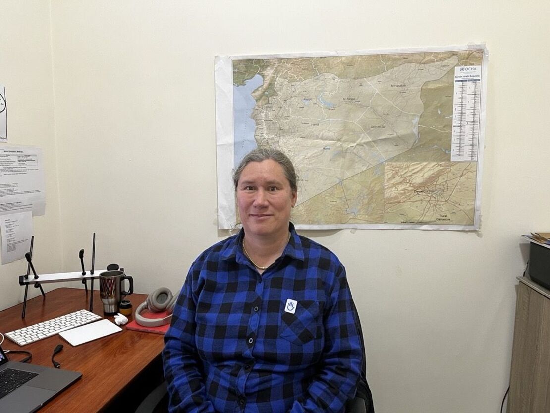 headshot of a woman wearing a blue and black plaid long-sleeved, sitting at a desk in front of a map on a beige wall. 