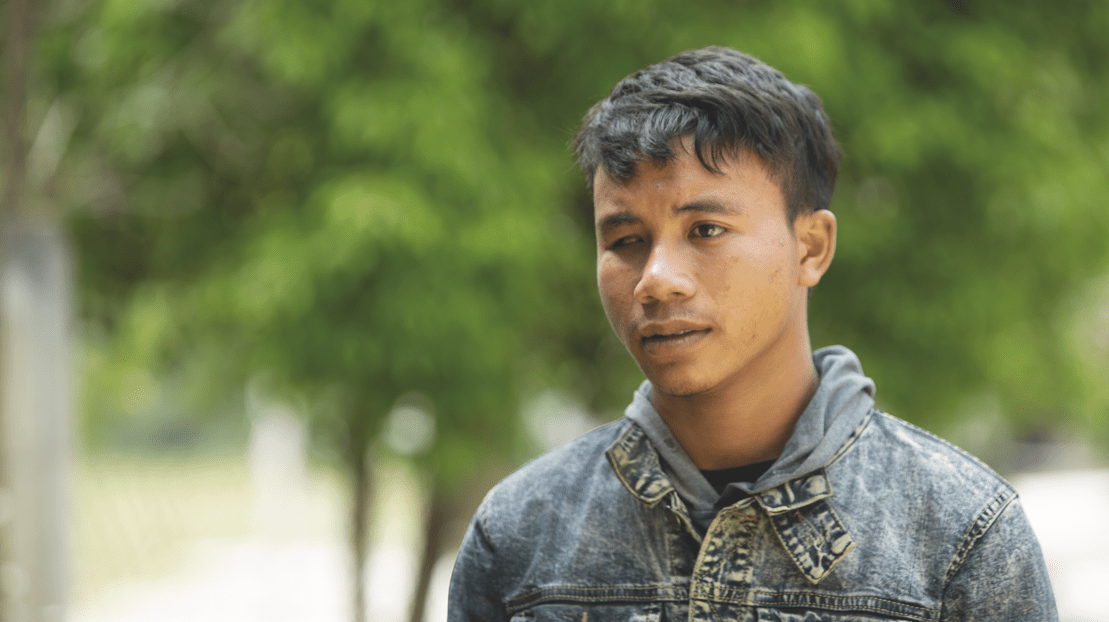 Portrait of Lithsouda, a young man missing an eye, wearing a denim jacket and looking into the distance on a background of green nature