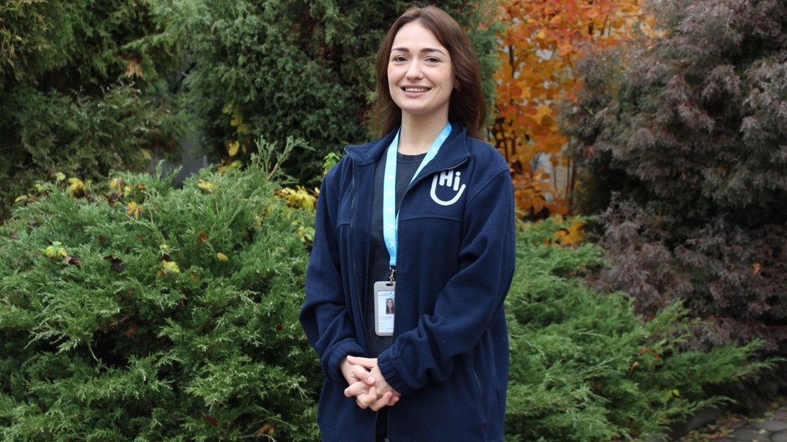 A woman is standing outdoors, surrounded by dense vegetation consisting of green shrubs and trees with autumn leaves. She is wearing a badge around her neck and a dark blue top with a light-coloured logo on the chest, and is holding her hands clasped in front of her.