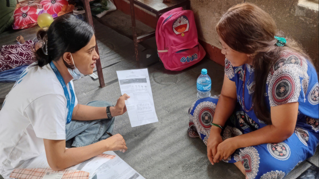 Two women are sitting in a room, facing each other; one is showing the other a sheet of paper whilst explaining something.