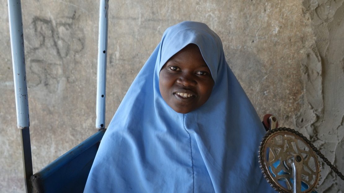 Portrait of Roumanatou, a Black woman wearing a blue headscarf, looking at the camera and smiling. 