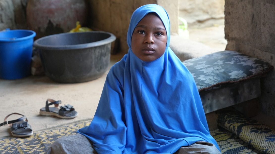 A little girl sits wearing a blue head covering staring directly facing the camera.