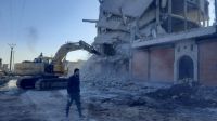 A man walks in front of an excavator woking on a collapsed building