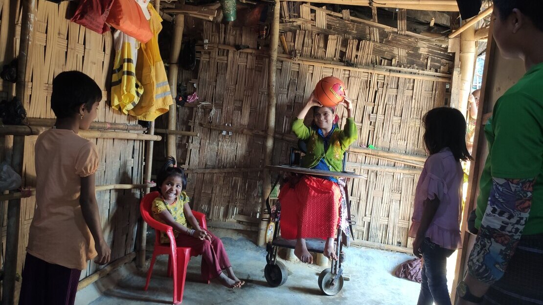 Group of four children playing with a basketball. In the center, a girl seated in a wheelchair holds the basketball over her head, smiling. 