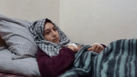 Teenage girl wearing a head scarf, laying down with a blanket on top of her. She looks solemn and into the distance. 
