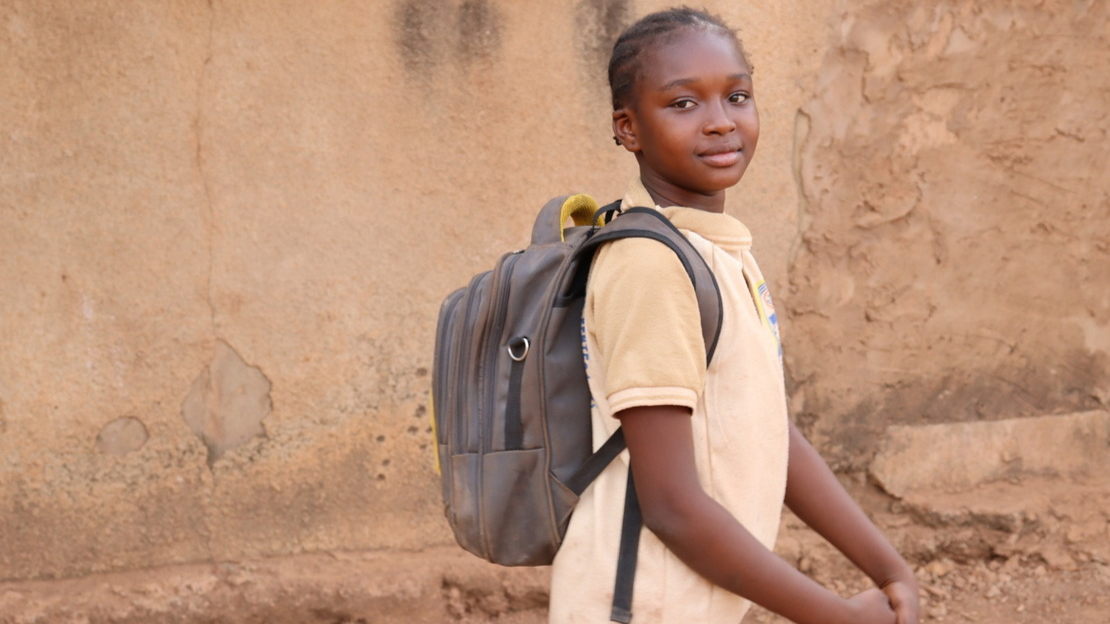 Portrait of Perpétue in profile, looking at the camera and smiling. She is carrying a backpack.