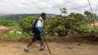 Raphaël walking using his walking devices on a dirt trail beside vegetation to Selembao inclusive school in the Democratic Republic of Congo. 