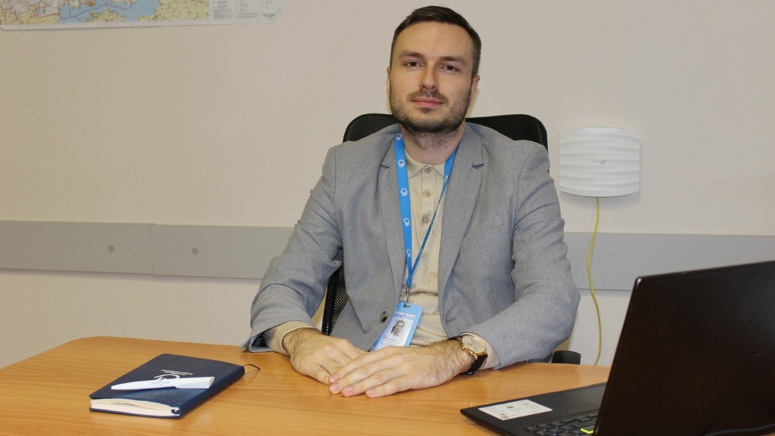 A person is sitting at a desk in an office setting. They are wearing a light-colored jacket and a name tag on a blue lanyard with a visible logo. Their hands are resting on the desk, which holds a notebook and an open laptop. In the background is a light-colored wall with a map hanging on it and a wall-mounted device.