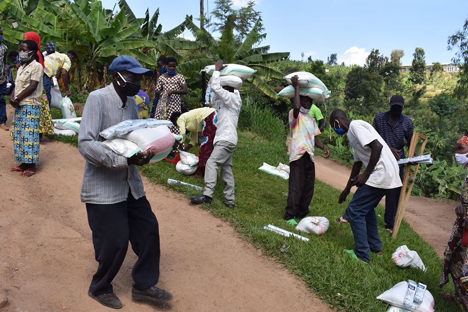 A dozen people wearing masks carrying and sorting through bags in a green field next to a dirt road