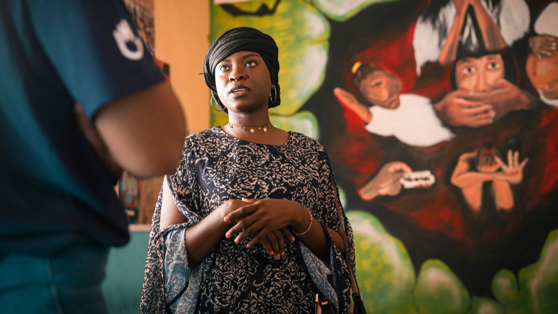 Awa stands looking at a person standing in front of her, whose t-shirt sleeve features a HI logo. Behind her, a wall with paintings of a young girl who hides her face with her hands, another who is pregnant, a third who is crying while two hands prevent her from speaking.