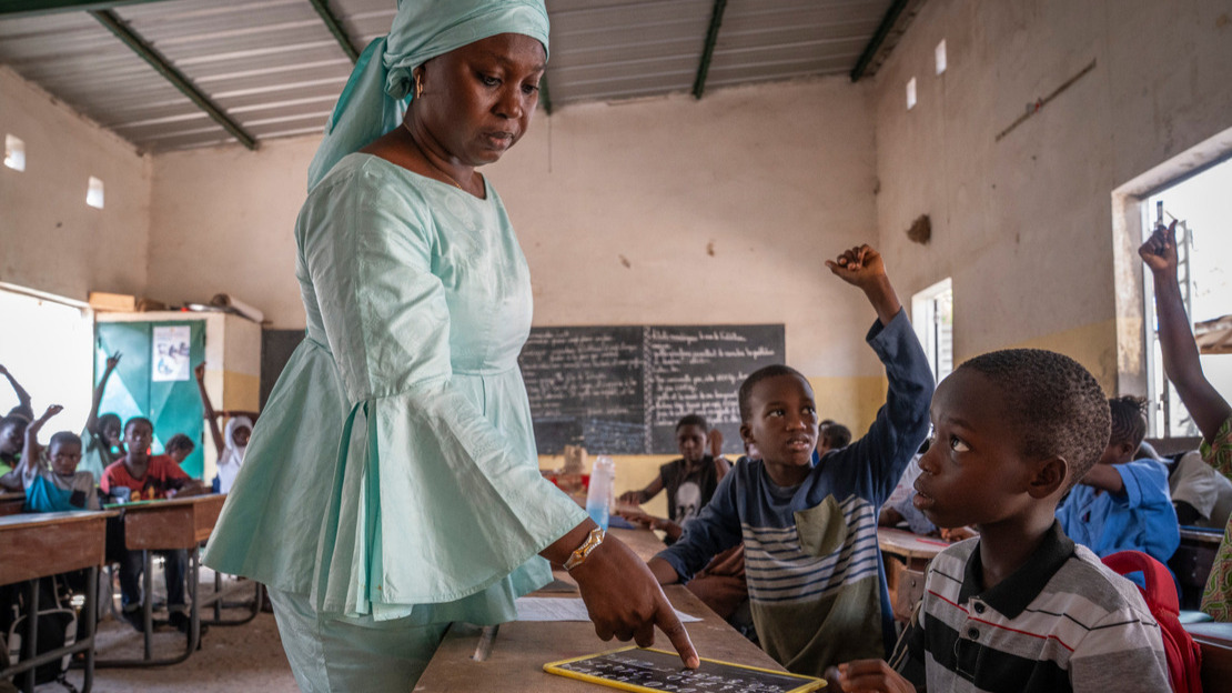 In a classroom, a woman is pointing to something on the blackboard of a boy sitting in the foreground.
