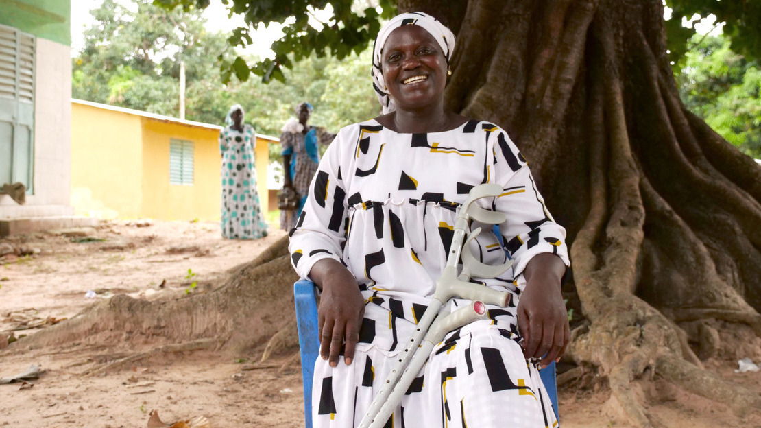 A woman sits on a blue plastic chair in front of a large tree. Dressed in a white dress with black and yellow patterns, she smiles at the camera. Crutches rest on her legs.