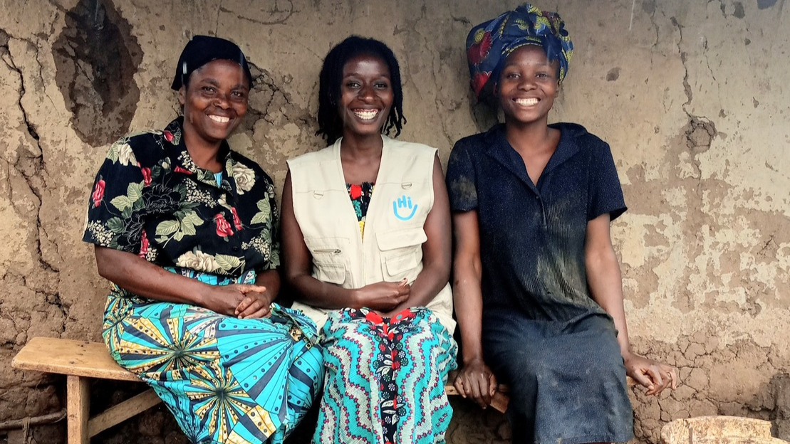 Three Black women sit on a bench. They are a mother and daughter with a mental health specialist. The three women are smiling.