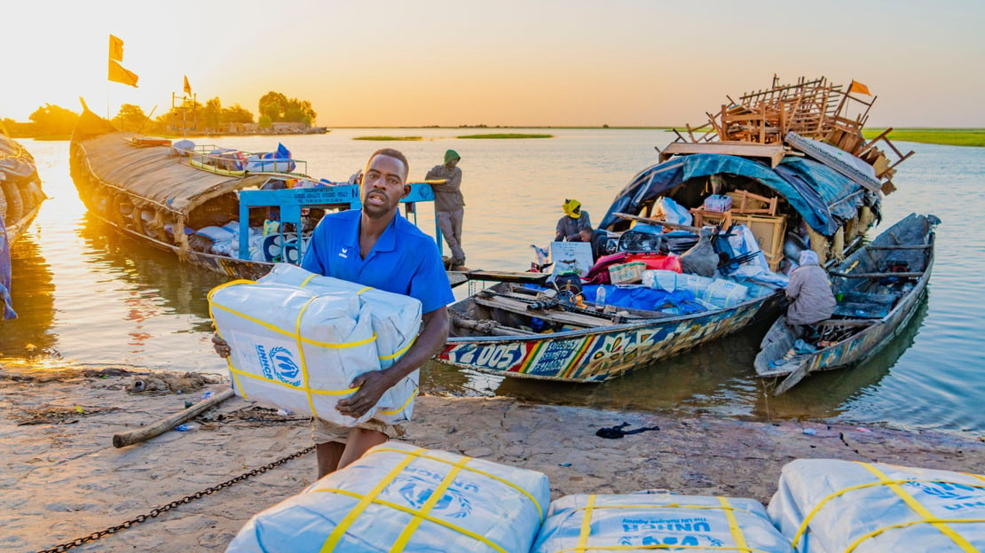 In the foreground, bags bearing the UNHCR logo. Behind, a man carries two bags in front of him, which he has just unloaded from two boats floating in the background.