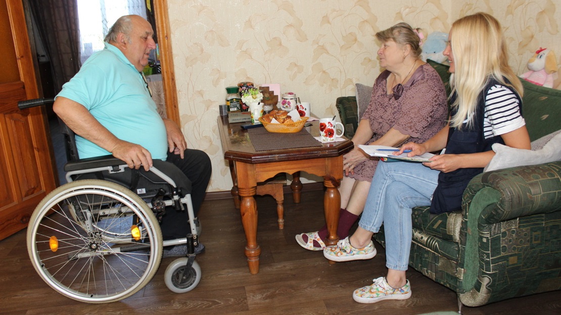 Three people are around a table; one man is in a wheelchair. They are chatting.