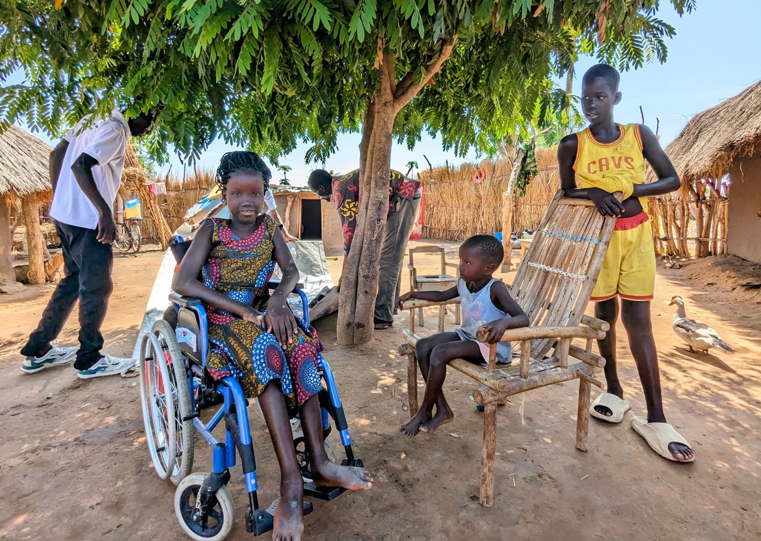 A little girl in a wheelchair in her classroom with her pupils and teacher standing next to her.