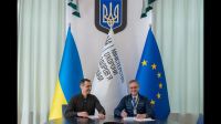 Two men sit smiling with pens at a desk with pens and paper in front of the Ukrainian flag, Ukrainian trident symbol 