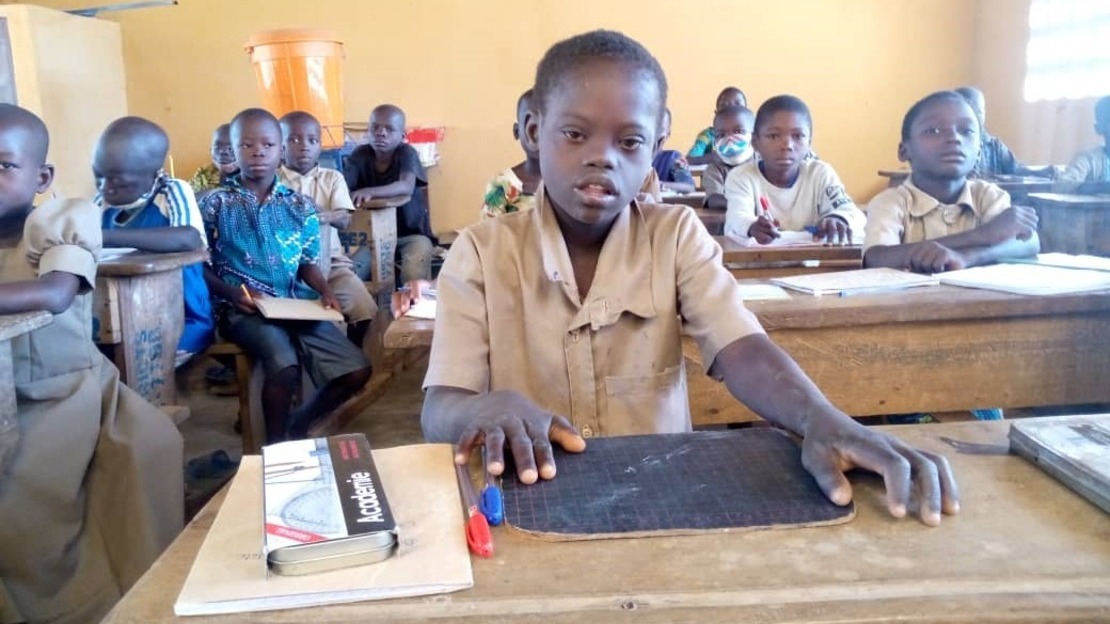 A young boy in a classroom amongst peers sitting on a school bench with his supplies in front of him.