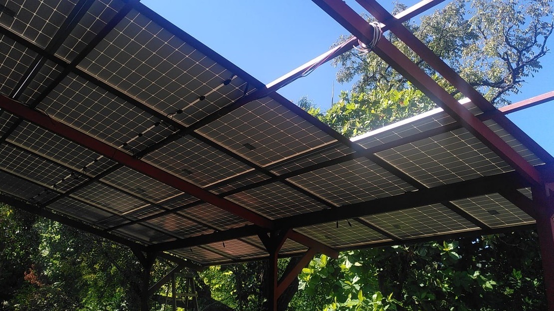 Low-angle view of a metal structure almost entirely covered with solar panels. Trees and blue sky can be seen through the empty spaces.