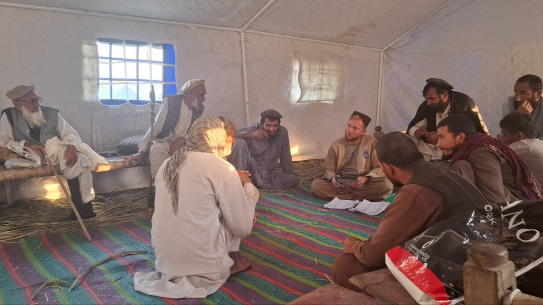 Under a canvas tent, a group of men are sitting in a circle on mats and benches. They are talking to a man wearing a jacket with HI printed on it.