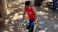 A young girl wearing a red t-shirt and jeans, smiles as she uses her new yellow walking frame