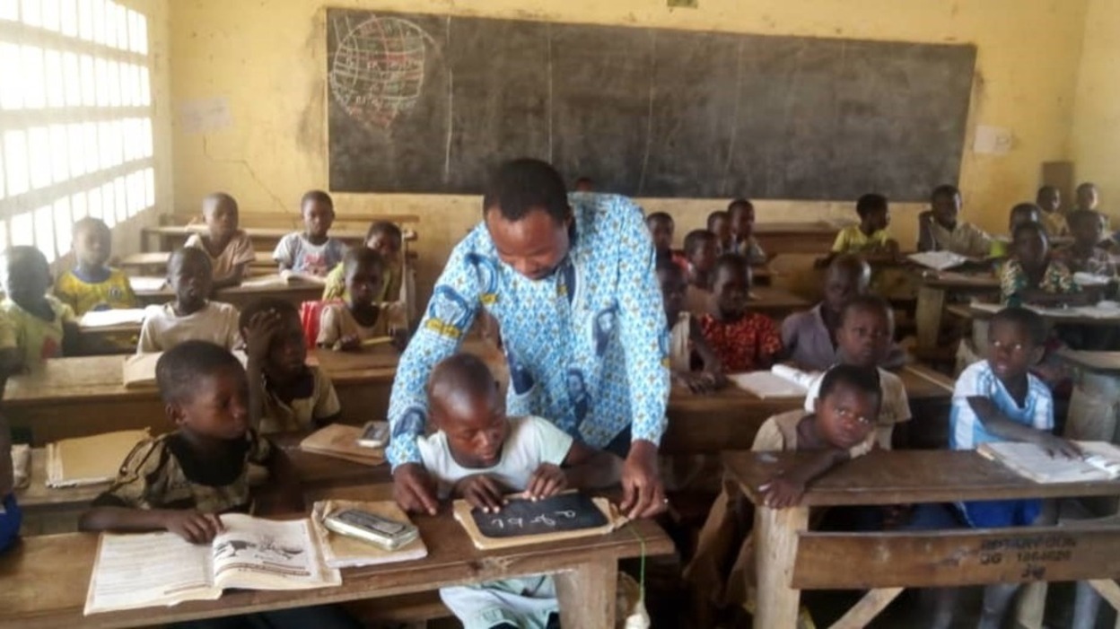 man leaning over a child sitting at a desk using a small chalkboard 