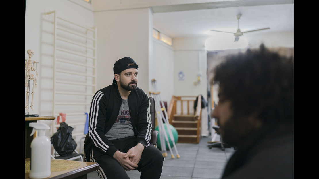 Man wearing a baseball hat and track suit sits on a bench in a physical therapy suite